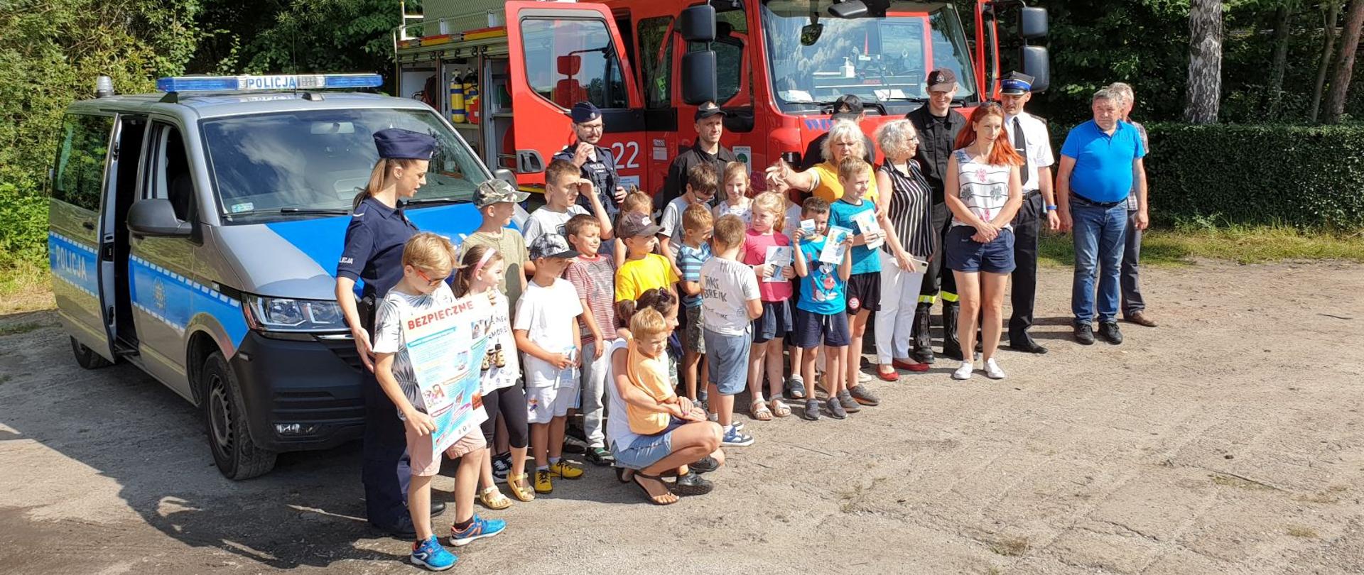 Zdjęcie przestawia uczestników Akcji Bezpieczne Wakacje na Grajdołku na tle samochodów Policji oraz Straży Pożarnej.