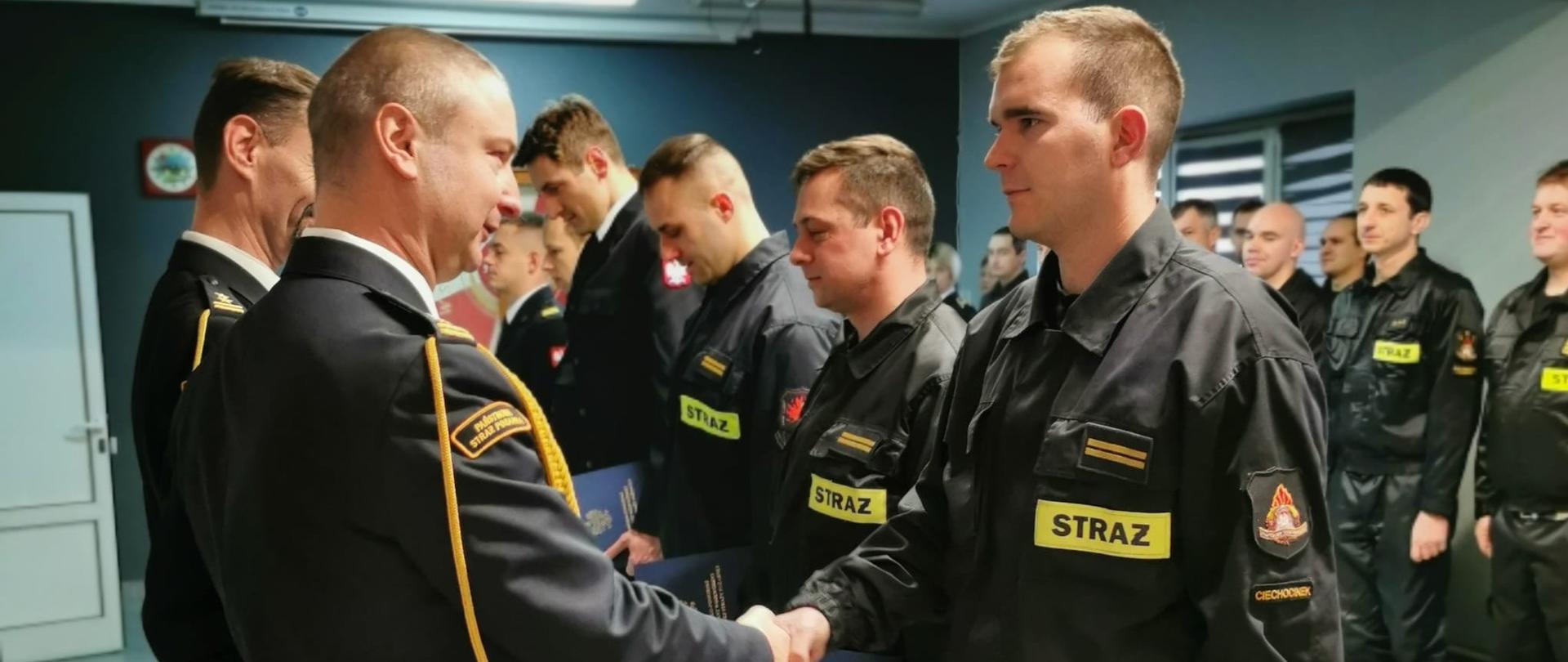 Strażacy w umundurowaniu koszarowym podczas wręczania przez komendanta powiatowego i Jego zastępcę aktów nadania wyższych stopni służbowych podczas uroczystej zbiórki z okazji Narodowego Święta Niepodległości