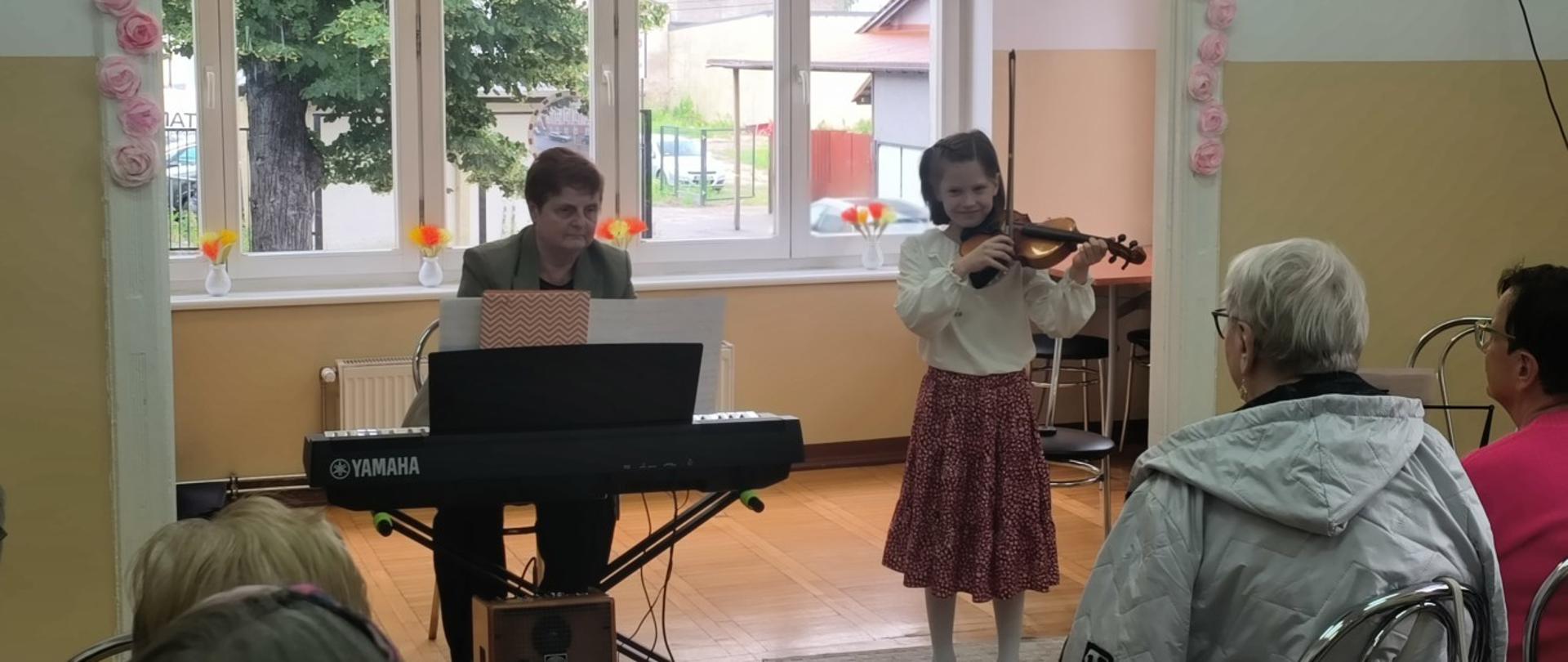 Zdjęcie kolorowe. W pomieszczeniu młoda dziewczynka gra na skrzypcach. Obok niej siedzi kobieta grająca na pianinie Yamaha. Z przodu widać głowy publiczności. Pomieszczenie ma duże okna, a na parapecie stoją wazony z kwiatami. Na ścianach papierowe ozdoby.