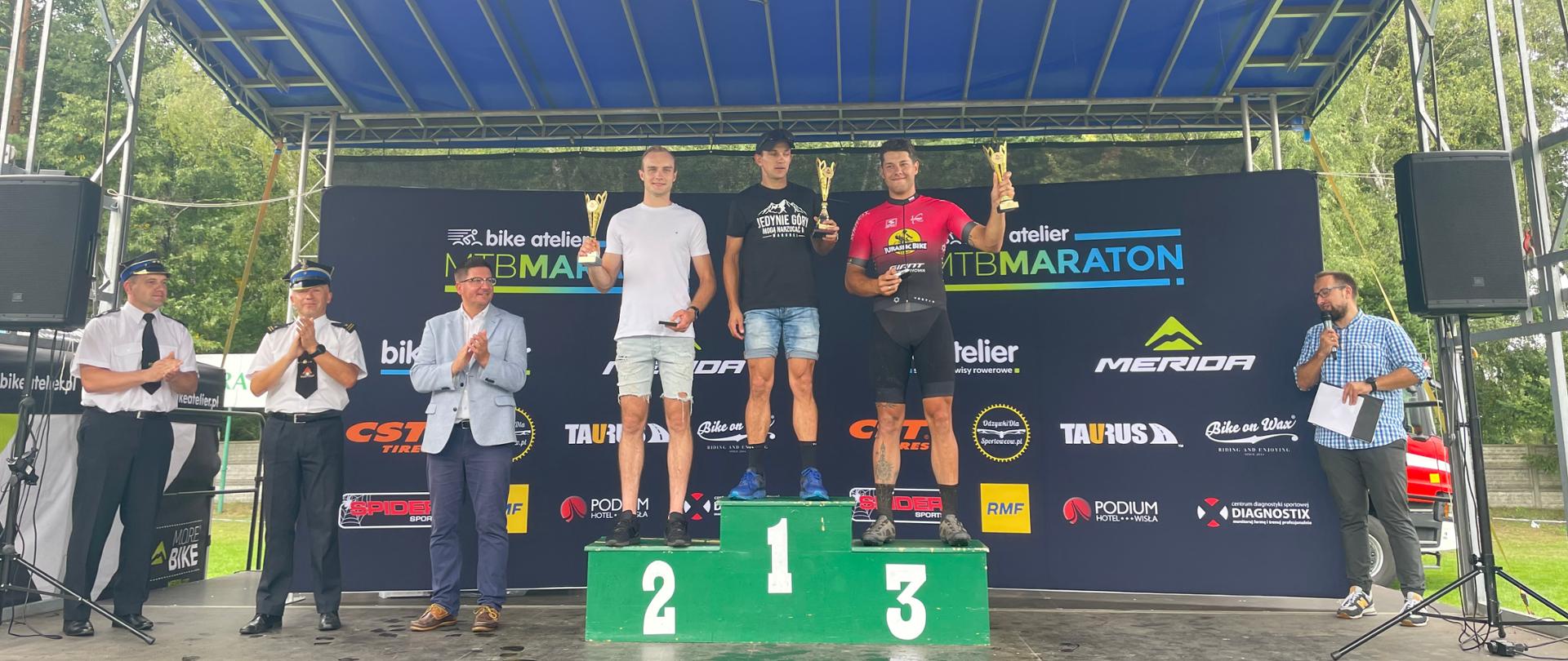 Zdjęcie przedstawia stojących na zadaszonej scenie na podium trzech uczestników maratony MTB z PSP. Na podium nr 1 stoi strażak ubrany w czarna koszulkę i dżinsowe spodenki i trzyma w lewej ręce podniesiony puchar. Na podium drugim stoi strażak ubrany w białą koszulkę i jasne spodenki i w prawej dłoni trzyma puchar. Na trzecim miejscu podium stoi strażak ubrany w czarno czerwoną koszulkę i czarne spodenki w lewej dłoni trzyma puchar. Po lewej stronie stoją dwaj strażacy ubrani w białe koszule wyjściowe i spodnie na głowie maja czapki wyjściowe obok nich stoi osoba ubrana w jasna marynarkę, niebieskie spodnie. Po prawej stronie podium stoi osoba ubrana w koszule w kratkę i ciemne spodnie w prawej ręce trzyma mikrofon, a w lewej białą kartkę. Za podium stoi wielki baner z napisem sponsorów