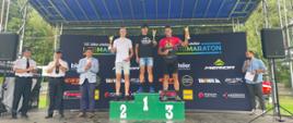 Zdjęcie przedstawia stojących na zadaszonej scenie na podium trzech uczestników maratony MTB z PSP. Na podium nr 1 stoi strażak ubrany w czarna koszulkę i dżinsowe spodenki i trzyma w lewej ręce podniesiony puchar. Na podium drugim stoi strażak ubrany w białą koszulkę i jasne spodenki i w prawej dłoni trzyma puchar. Na trzecim miejscu podium stoi strażak ubrany w czarno czerwoną koszulkę i czarne spodenki w lewej dłoni trzyma puchar. Po lewej stronie stoją dwaj strażacy ubrani w białe koszule wyjściowe i spodnie na głowie maja czapki wyjściowe obok nich stoi osoba ubrana w jasna marynarkę, niebieskie spodnie. Po prawej stronie podium stoi osoba ubrana w koszule w kratkę i ciemne spodnie w prawej ręce trzyma mikrofon, a w lewej białą kartkę. Za podium stoi wielki baner z napisem sponsorów 