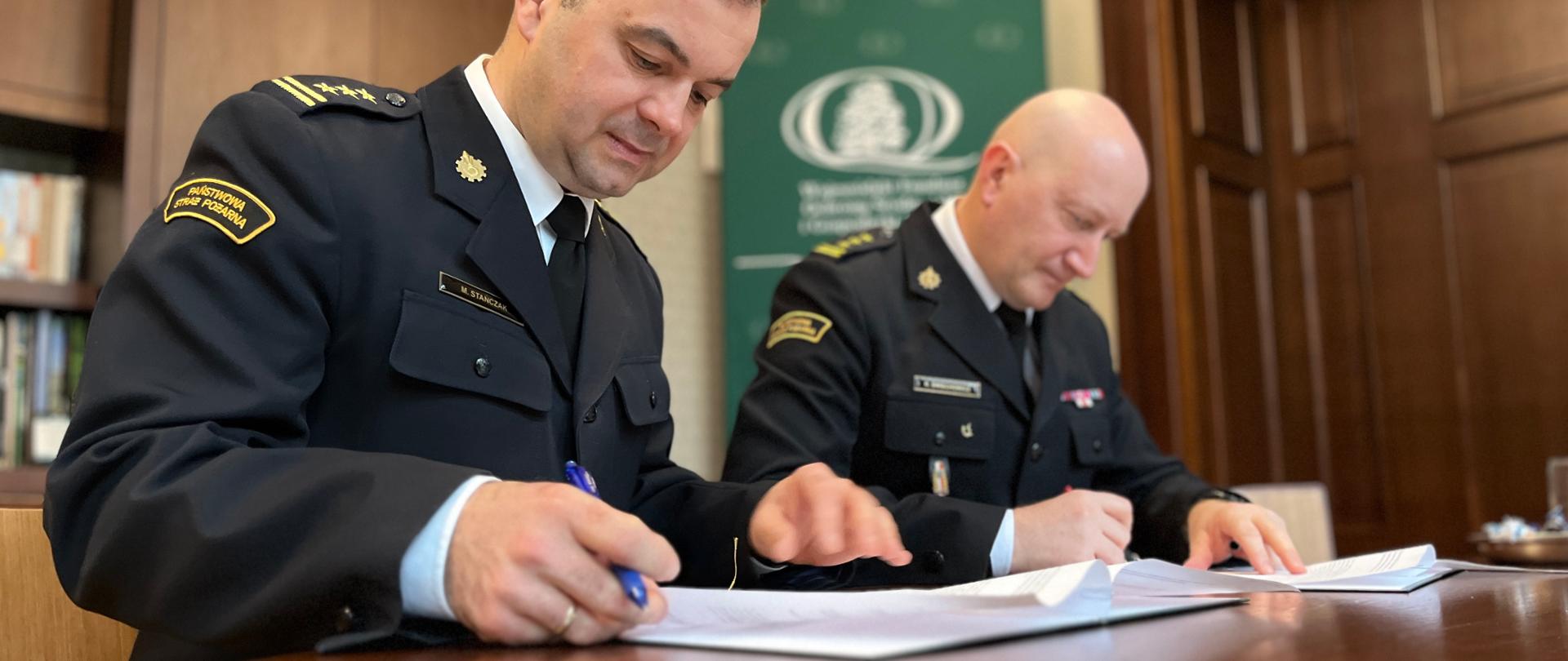 Dwóch strażaków, w mundurach wyjściowych, przy stole podpisuje dokumenty. W tle wystrój gabinetu.