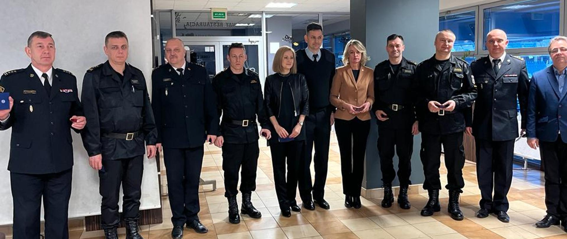 Grupa otrzymująca odznaczenie stojąca frontem w szeregu do osoby robiącej zdjęcie, pierwszy z lewej st. bryg. Janisław Buller Komendant Miejski PSP w Bydgoszczy. Wszyscy znajdują się w sali konferencyjnej, w tle widać przeszklenia ścienne sali. Podłoga w terakocie kremowej z brązowymi wstawieniami. 