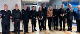Grupa otrzymująca odznaczenie stojąca frontem w szeregu do osoby robiącej zdjęcie, pierwszy z lewej st. bryg. Janisław Buller Komendant Miejski PSP w Bydgoszczy. Wszyscy znajdują się w sali konferencyjnej, w tle widać przeszklenia ścienne sali. Podłoga w terakocie kremowej z brązowymi wstawieniami. 