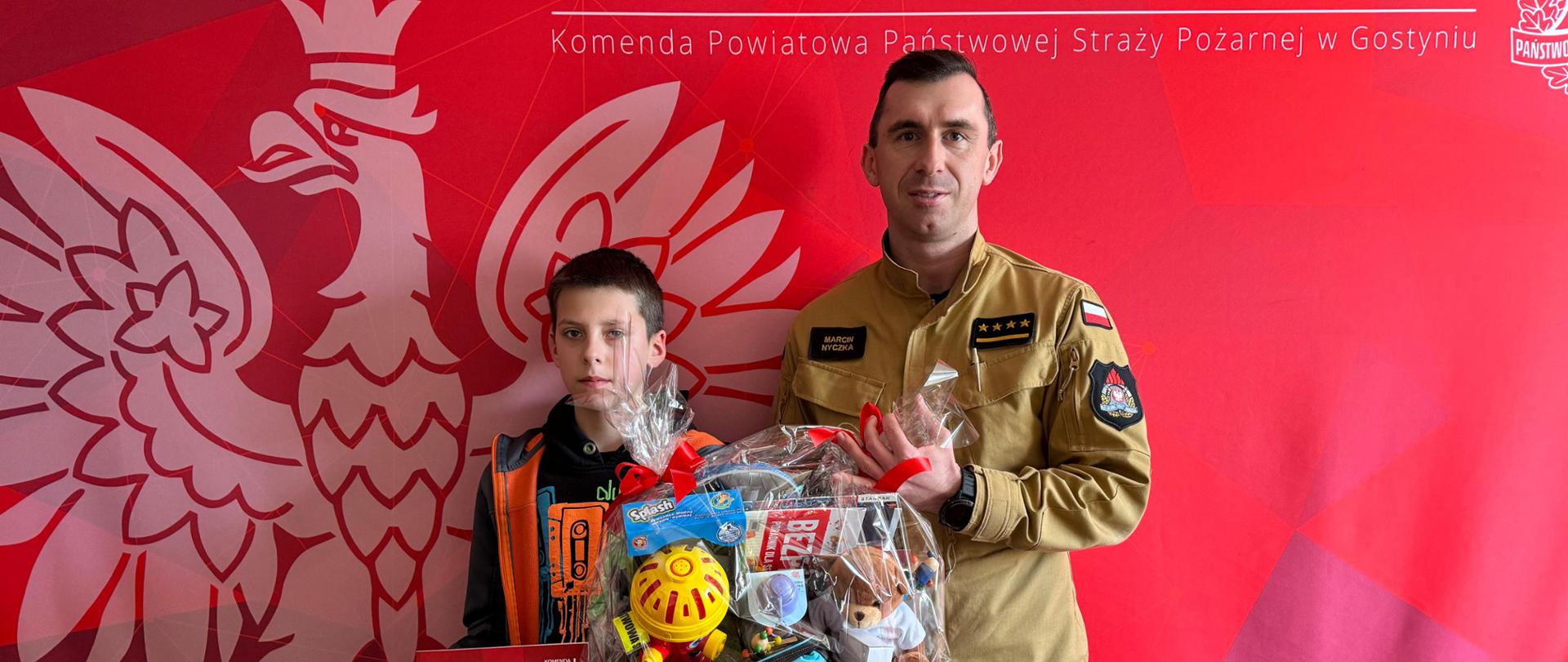 Chłopca Daniela oraz komendanta stojących obok siebie i pozujących do zdjęcia. Chłopiec trzyma w ręce upominki oraz list otrzymane od komendanta.