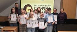 Na zdjęciu grupa uczniów biorących udział w konkursie wiedzy o Fryderyku Chopinie. Wszyscy uczniowie są uśmiechnięci i trzymają dyplomy. W tle na interaktywnej tablicy wizualizacja Fryderyka Chopina oraz napis "świat dźwięków Chopina konkurs". Po prawej i lewej stronie obrazka ławki szkolne z krzesełkami.