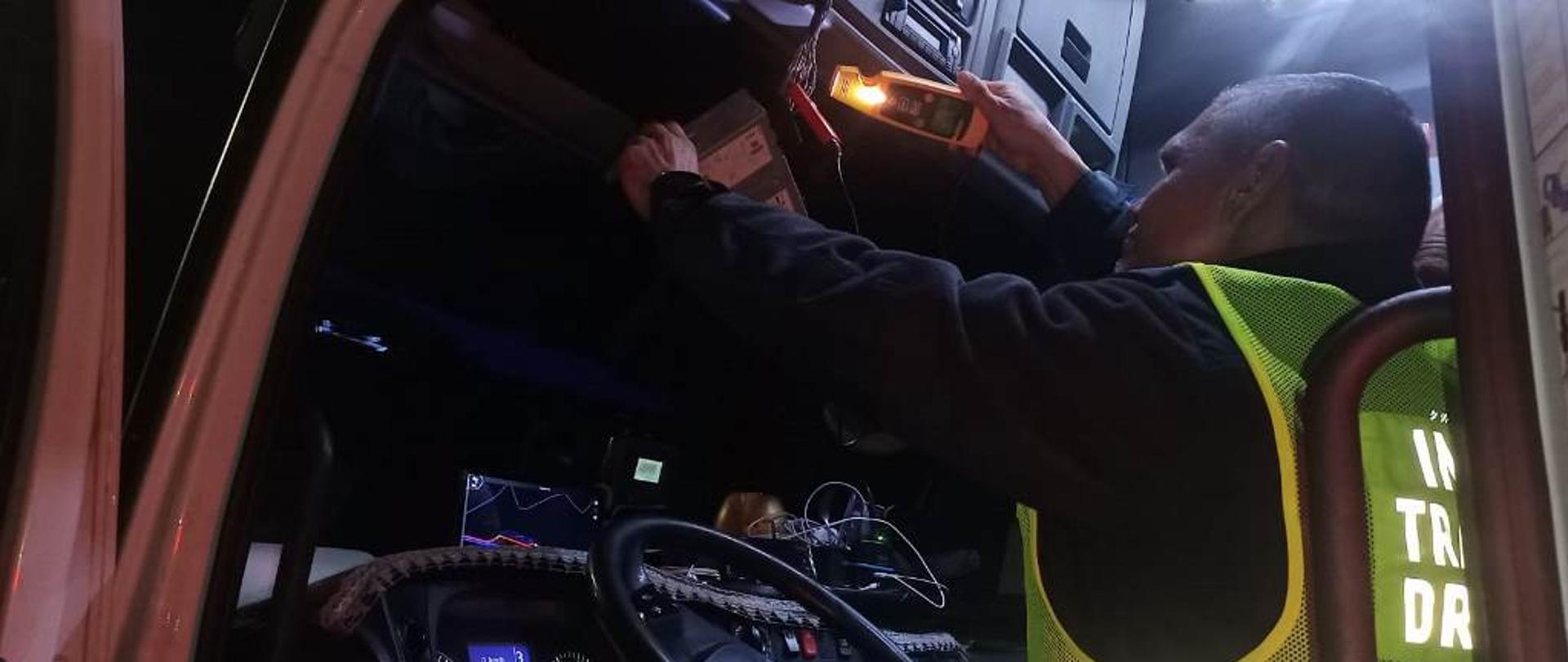 Umundurowany inspektor lubuskiej Inspekcji Transportu Drogowego siedzi na fotelu kierowcy w kontrolowanej ciężarówce. Funkcjonariusz podłącza specjalistyczny przyrząd kontrolno-pomiarowy do instalacji tachografu zamontowanego w pojeździe.