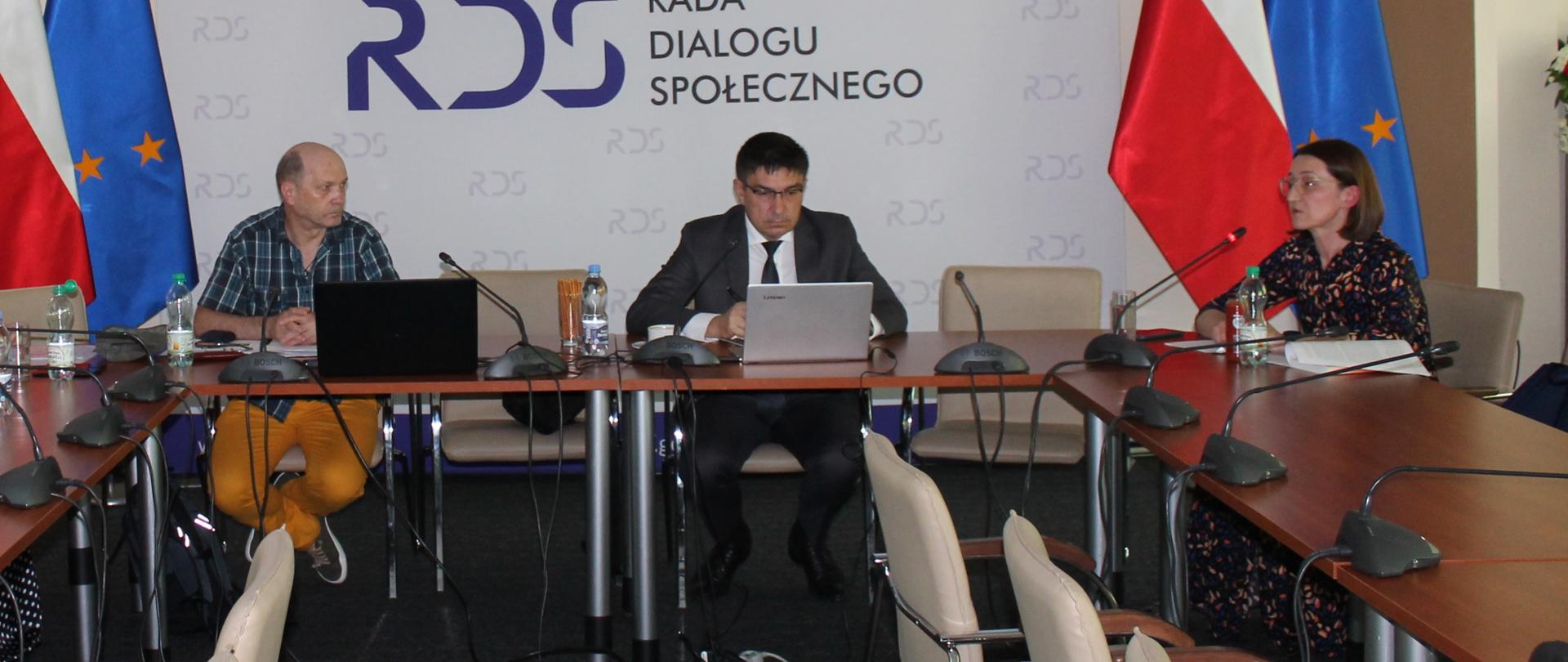 Na zdjęciu stół prezydialny. Od lewej strony siedzi przedstawiciel związków zawodowych, następnie Przewodniczący Zespołu Wojciech Ilnicki oraz Agata Olkińska - zastępca Dyrektora Departamentu Dialogu i Partnerstwa Społecznego. W tle siedzących baner z napisem Rada Dialogu Społecznego.