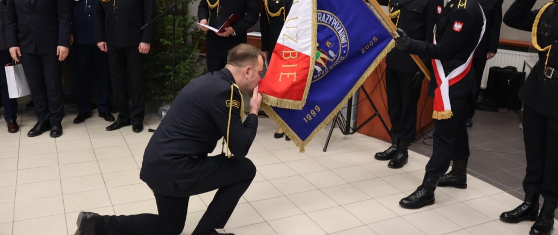 Zdjęcie wykonane w Sali tradycji komendy miejskiej Państwowej Straży Pożarnej podczas uroczystej zbiórki dotyczącej zdania obowiązków komendanta miejskiego. Na zdjęciu widać moment pożegnania ze sztandarem komendanta miejskiego Państwowej Straży Pożarnej w Radomiu. Funkcjonariusz klęczy na jednym kolanie przed pochylonym sztandarem. Jedną ręką trzyma róg sztandaru składając na niego pocałunek. Pochylony sztandar w rękach trzyma jeden z trzech funkcjonariuszy tworzących poczet sztandarowy. 