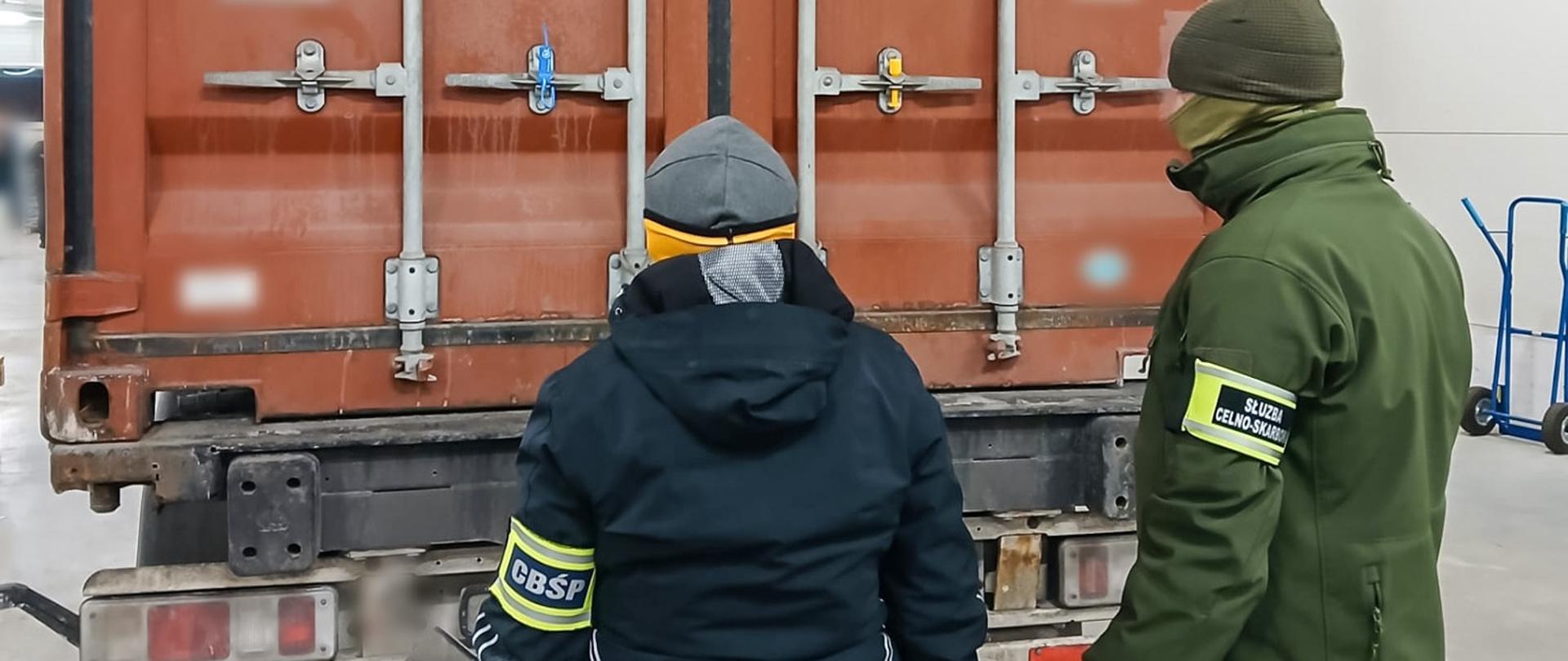 Dwie osoby stoją przed zamkniętymi drzwiami kontenera; widoczne są metalowe zamki i zabezpieczenia drzwi.