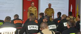 Szkolenie podstawowe strażaka ratownika OSP. Zdjęcie przedstawia świetlicę Komendy Powiatowej PSP w Rawiczu. Przy stołach ustawionych w rzędy, siedzą uczestniczki i uczestnicy szkolenia.Część z nich ma na sobie mundury koszarowe w kolorze czarnym, część koszulki w kolorze czarnym. Rozwiązują test egzaminacyjny. Przed nimi stoi dwóch strażaków PSP w ubraniach służbowych koloru piaskowego, członków komisji egzaminacyjnej. Na ekranach wyświetlany jest test egzaminacyjny.