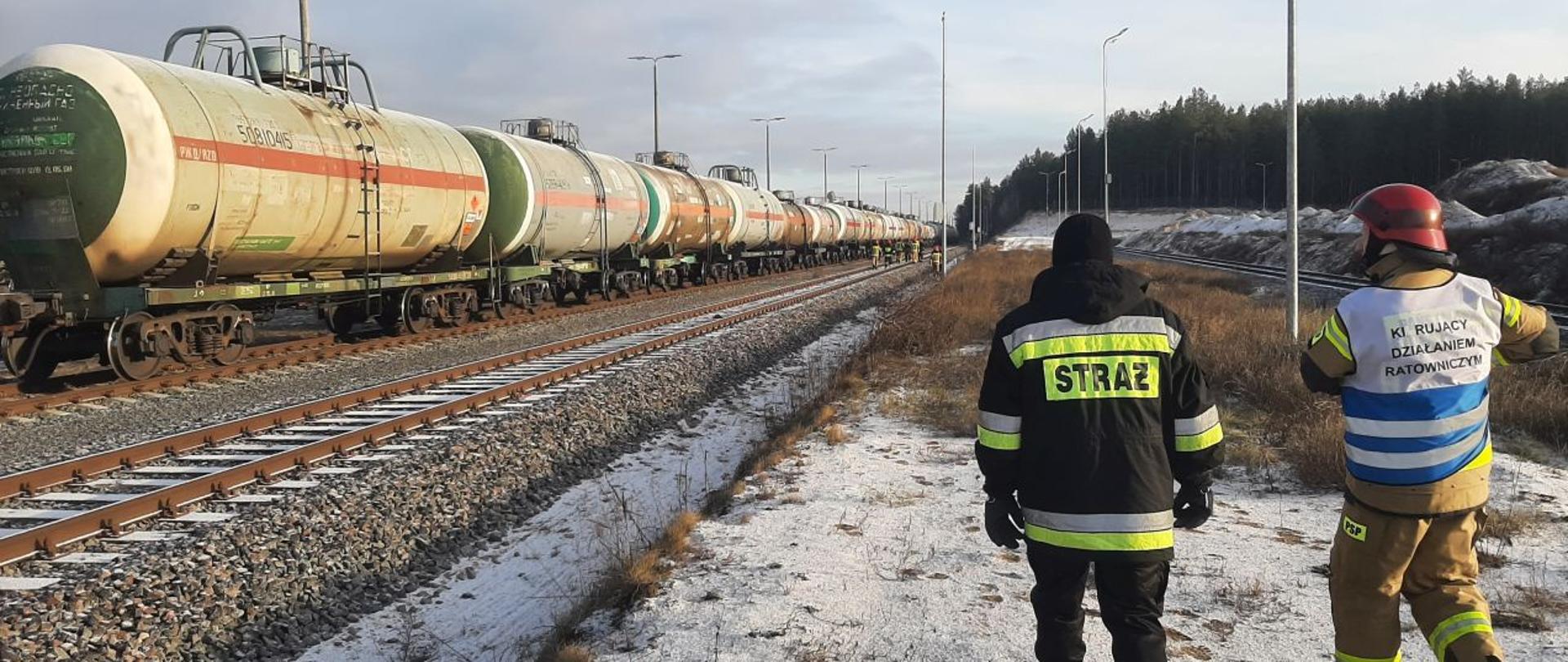 Widoczny skład kolejowy z cysternami, w tle strażacy pracujący przy uszkodzonym zaworze, widoczny dowódca akcji oraz oficer operacyjny