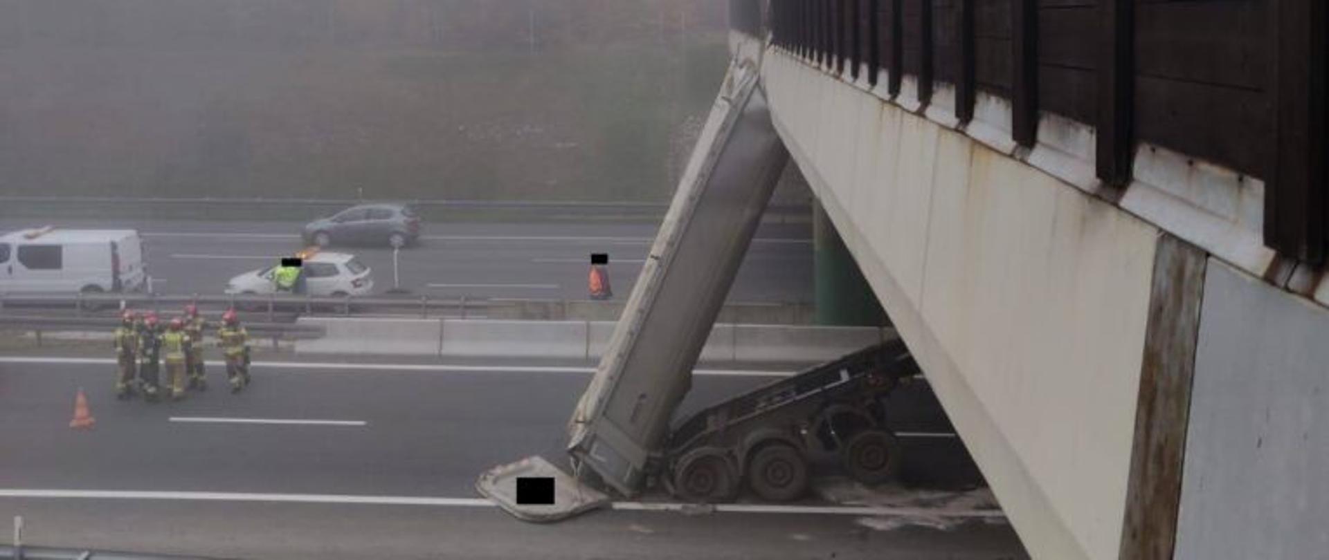 Zdjęcie przedstawia naczepę ciężarówki, która nie opuszczona uderzyła w wiadukt na autostradzie A4 - centralny element kadru zdjęcia. W lewej części kadru stoi grupa strażaków w mundurach specjalnych w hełmach koloru czerwonego na pasie autostrady. Na przeciwnym pasie drogi stoją trzy samochody osobowe oraz dwie osoby w odblaskowych ubraniach. Na drugim planie zdjęcia widoczny las we mgle.