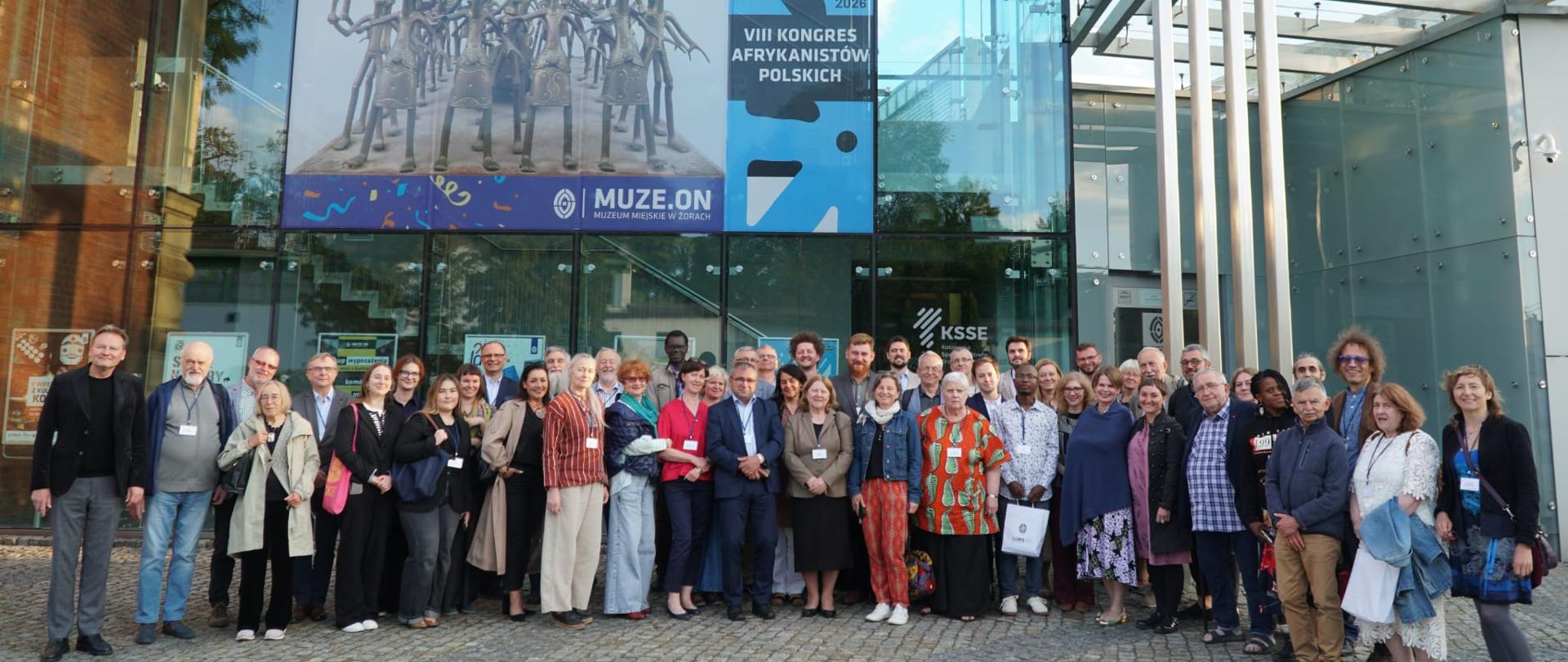 Group photo of 48 participants of the 8th Africanist Congress in Żory; some are wearing passes; almost all are smiling for the photo; in the background is the Żory Museum building; a modern glass facade; the building is two stories high; posters hang on the windows; the largest poster announces that the Żory Museum is celebrating its 25th anniversary; a smaller poster announces the 8th Africanist Congress in Żory; smaller posters displayed on the ground floor announce upcoming events; the area in front of the Museum is paved with gray cobblestones; decorative elements are paved with darker gray cubes