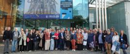 Group photo of 48 participants of the 8th Africanist Congress in Żory; some are wearing passes; almost all are smiling for the photo; in the background is the Żory Museum building; a modern glass facade; the building is two stories high; posters hang on the windows; the largest poster announces that the Żory Museum is celebrating its 25th anniversary; a smaller poster announces the 8th Africanist Congress in Żory; smaller posters displayed on the ground floor announce upcoming events; the area in front of the Museum is paved with gray cobblestones; decorative elements are paved with darker gray cubes