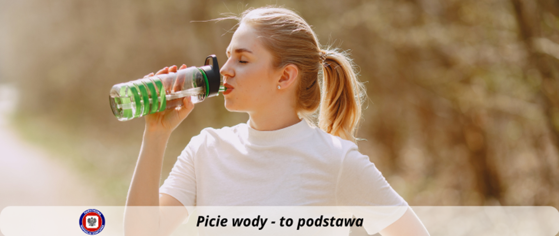 Młoda blondynka w białej koszulce pije wodę z bidonu. Poniżej na jasnym półprzezroczystym pasku ciemny napis Picie wody - to podstawa. Po jego lewej stronie logo Państwowej Inspekcji Sanitarnej.