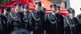 Szereg strażaków w ciemnych mundurach galowych i czapkach rogatywkach stoi na baczność podczas oficjalnej uroczystości na świeżym powietrzu.