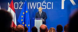 Wicepremier Jacek Sasin przemawia przy mównicy, obok flaga Unii i Austrii, w tle napis nowe możliwości. Na pierwszym planie nieostre głowy słuchaczy