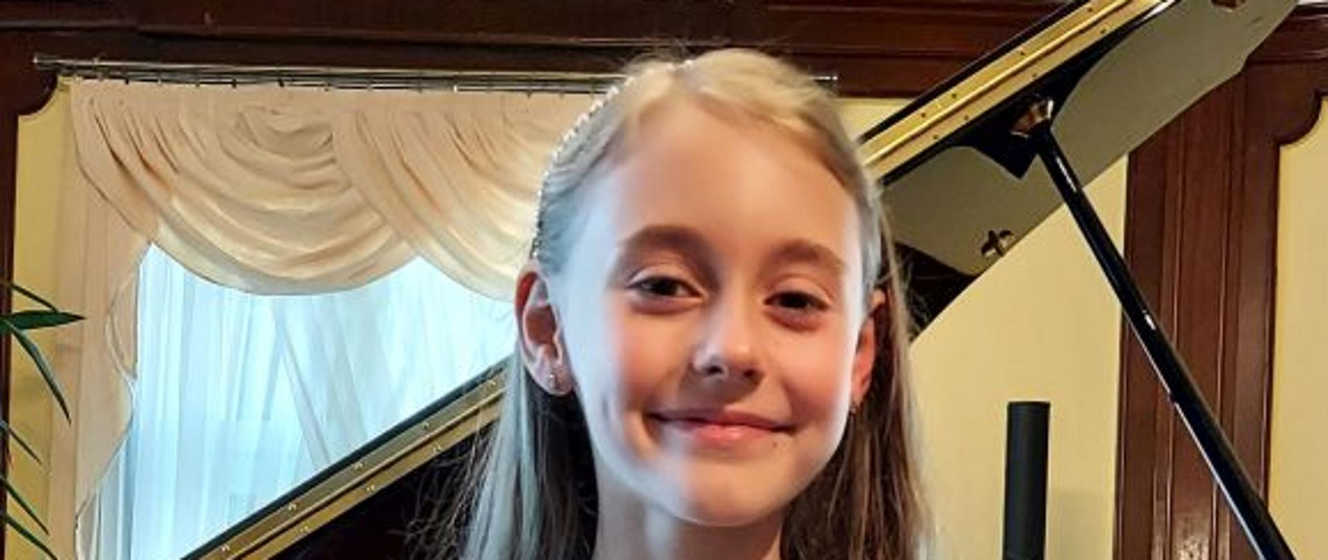 Alicja Puchalik, uczennica, siedzi na tle fortepianu w sali kameralnej Państwowej Szkoły Muzycznej I i II stopnia w Słupsku. Jest bardzo zadowolona,. Ttrzyma w rękach dyplom.