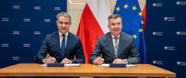 Za stolikiem siedzi minister Wieczorek i mężczyzna w szarym garniturze, obaj trzymają długopisy i podpisują leżące przed nimi dokumenty, za nimi pod ścianą flagi Polski i UE.