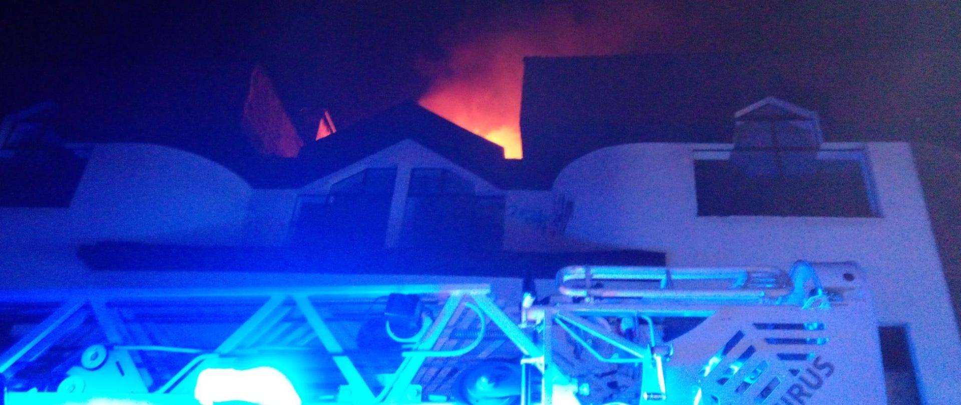 Zdjęcie przedstawia fragment drabiny mechanicznej stojącej na tle opuszczonego budynku gdzie doszło do pożaru. Nad dachem widać łunę bijąca od ognia.