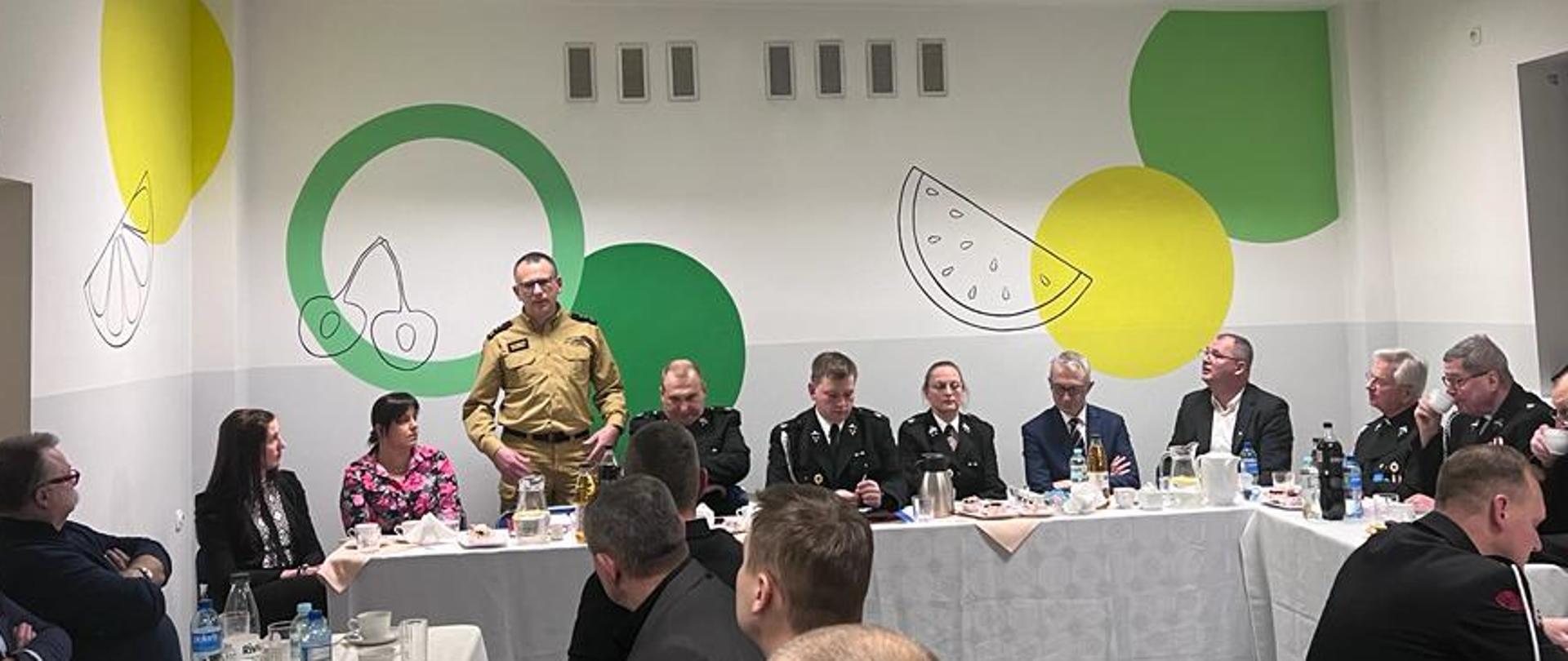 Grupa mężczyzn i kobiet siedzi w oświetlonej sali za stołem prezydialnym, jeden mężczyzna ubrany w mundur straży pożarnej przemawia na stojąco