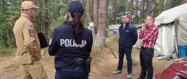 Na zdjęciu widać grupę pięciu osób stojących na terenie leśnego obozowiska harcerskiego. W centrum kadru znajduje się policjantka w ciemnym mundurze z napisem "POLICJA" na plecach, zwrócona tyłem do aparatu. Obok niej stoi strażak w beżowym mundurze z naszywkami służbowymi, rozmawiający z trzema cywilami. Dwóch z nich ubranych jest w ciemne ubrania, a trzeci – w czerwoną koszulę w kratę. Po prawej stronie częściowo widoczny jest czerwony samochód z tablicą rejestracyjną zaczynającą się od „ZSZ 82 WX”. W tle znajdują się namioty obozowe harcerzy i drzewa sosnowe. W lewym górnym rogu znajduje się logo polskiej policji. Zdjęcie dokumentuje wizytę służb na terenie obozu, prawdopodobnie w celu kontroli bezpieczeństwa.