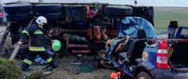 Na zdjęciu wraki pojazdu ciężarowego oraz samochodu osobowego w kolorze niebieskim oraz idącego strażaka w ubraniu specjalnym z hełmem koloru białego