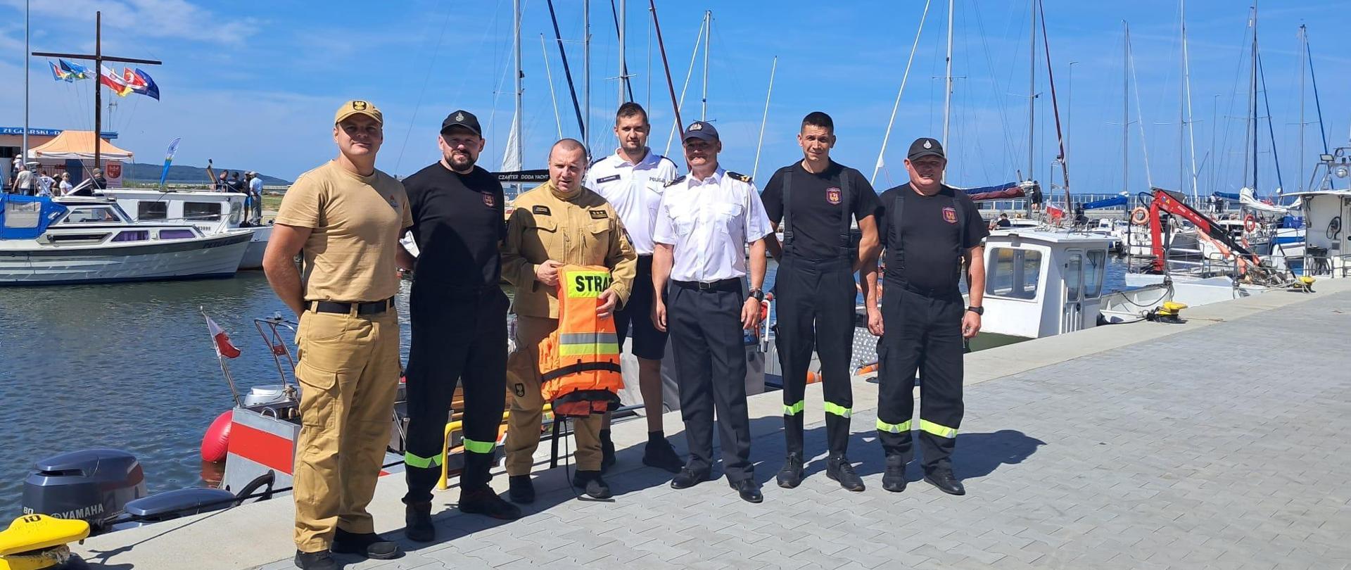 W porcie na brzegu stoi grupa mundurowych. Za mini w tle łodzie i jachty, niebieskie niebo, pogodnie.