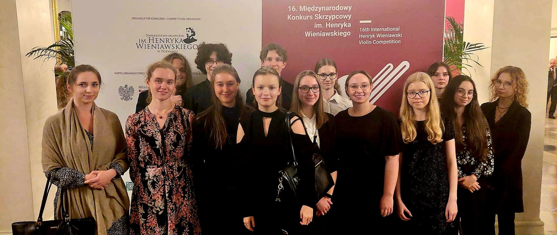Zdjęcie uczniów i nauczycielki w foyer Filharmonii Poznańskiej przy ściance reklamującej Wyjazd na przesłuchania 16. Międzynarodowy Konkurs Skrzypcowy im. H. Wieniawskiego