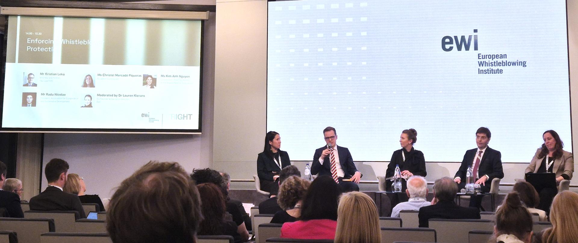 Pięć osób siedzi na scenie sali konferencyjnej na tle wyświetlacza z napisem "EWI - European Whistleblowing Instytite", kilkadziesiąt osób słucha wystąpień