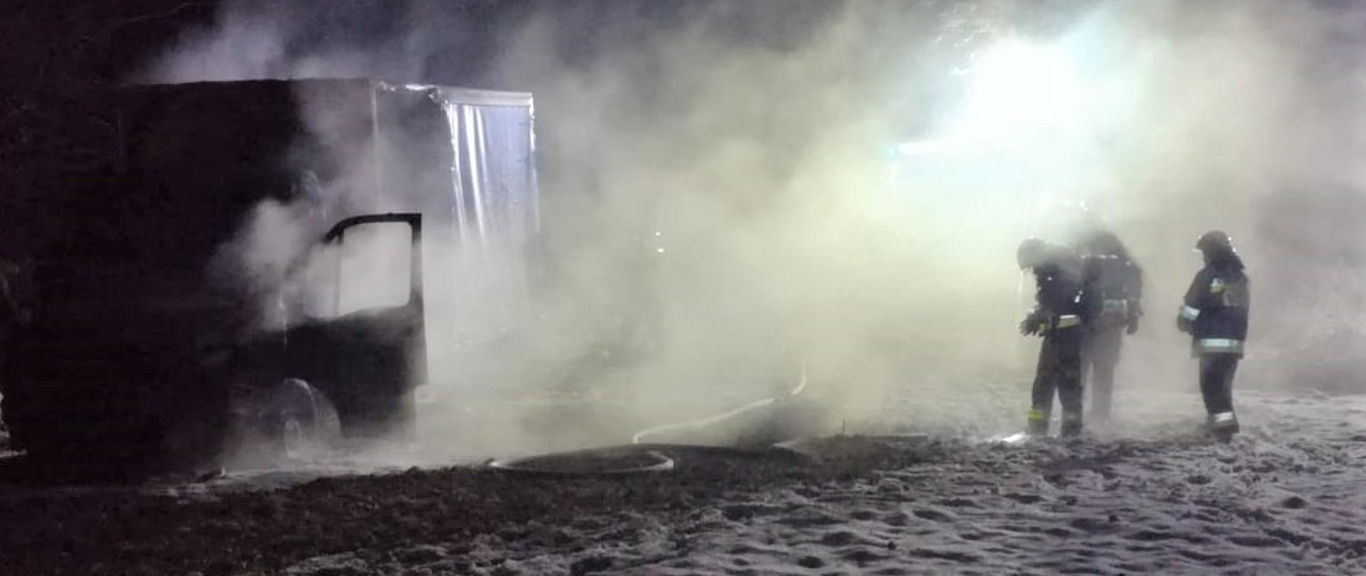 Na zdjęciu widzimy miejsce prowadzenia akcji ratowniczo-gaśniczej: po lewej stronie widać samochód po pożarze, z prawej stoją ratownicy. Wszystko osnute dymem i parą wodną.