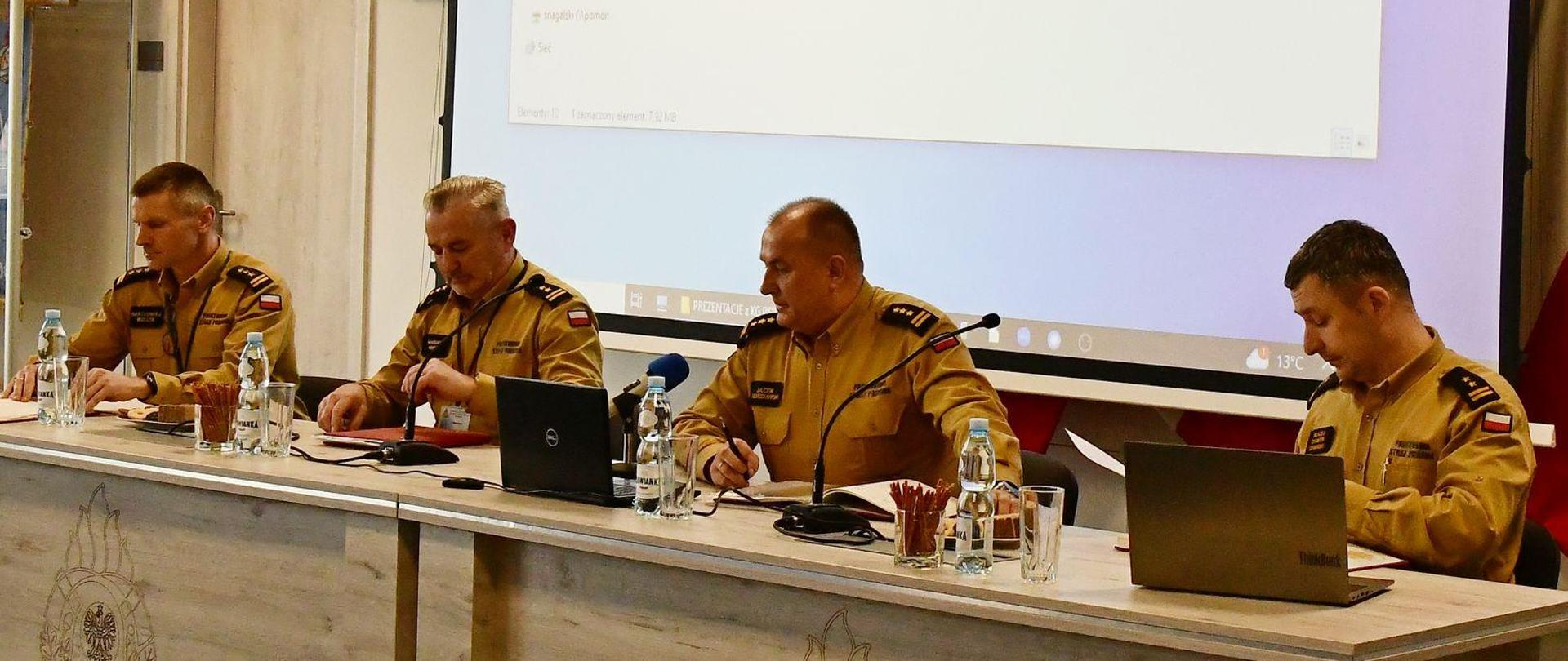 Trzech strażaków w mundurach koloru musztardowego siedzi za biurkami na których są laptopy paluszki woda oraz mikrofony na ekranie wyświetlana jest prezentacja.