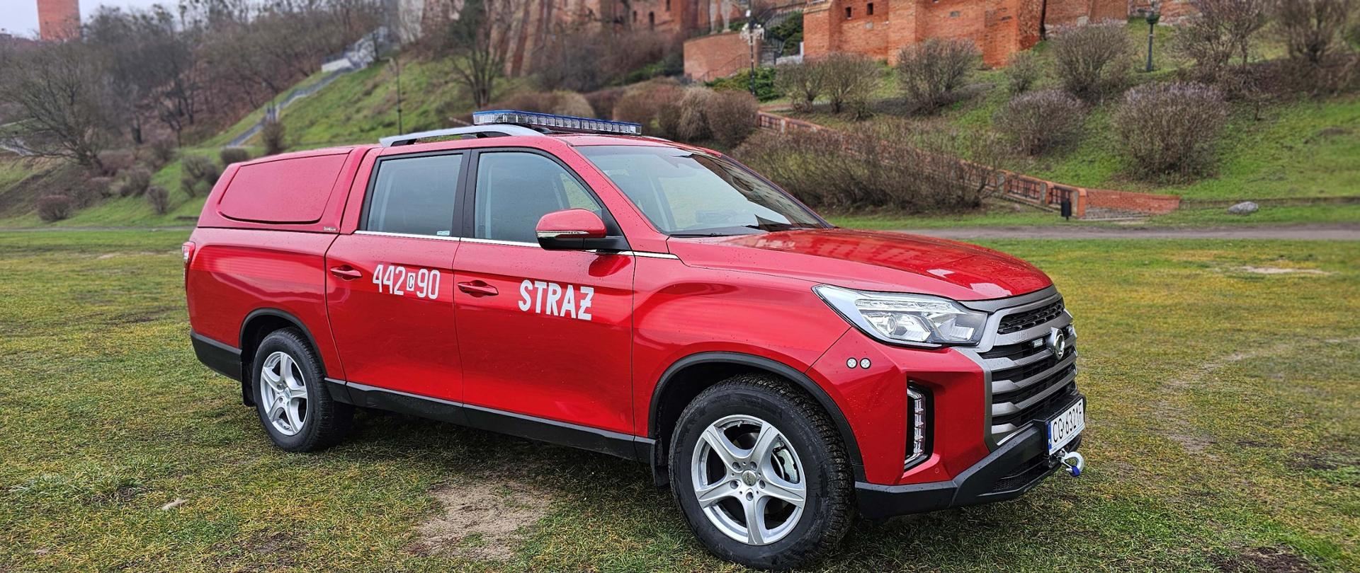 Czerwony strażacki samochód typu pick-up stojący na zielonym terenie. W tle gotycka zabudowa starego miasta.