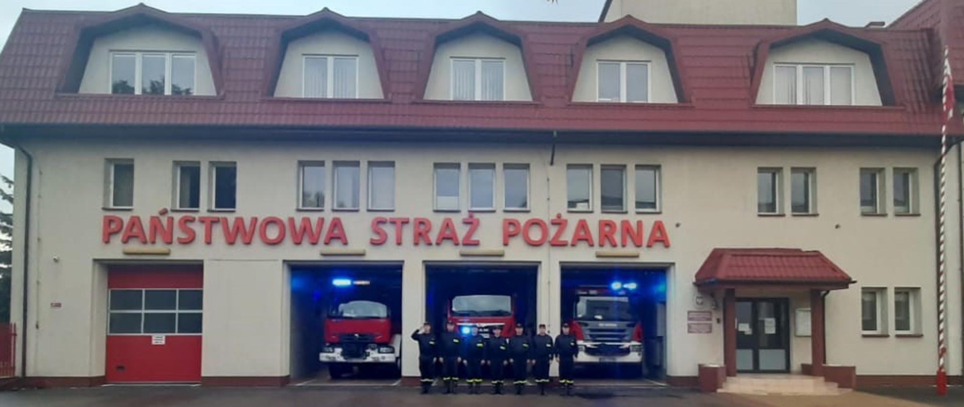 Strażacy z KP PSP w Bielsku Podlaskim ustawieni w szeregu przed budynkiem Komendy oddają hołd zmarłemu strażakowi z KM PSP w Grudziądzu. W samochodach ratowniczo-gaśniczych stojących za strażakami włączone są sygnały świetlne.