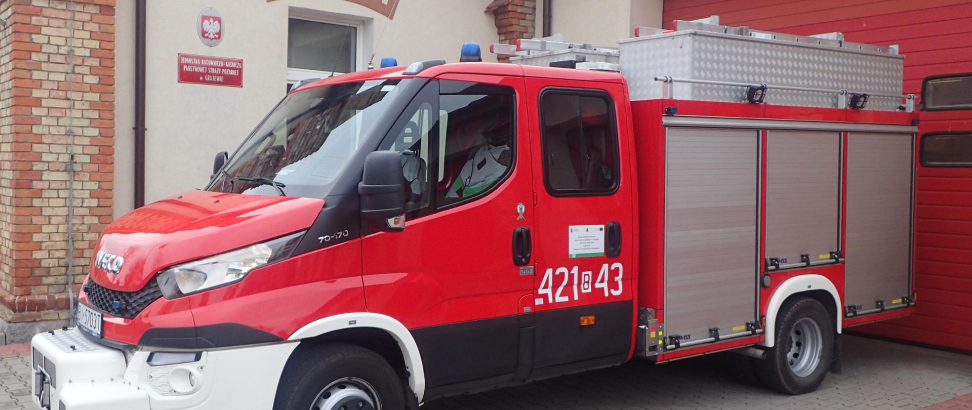 Na zdjęciu samochód lekki ratownictwa technicznego Iveco – SLRt w kolorze czerwonym z białym zderzakiem z przodu, boczne rolety w kolorze srebrnym. W tle budynek KP PSP w Grajewie 