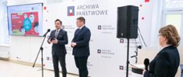 W białej sali pod ścianką z napisem Archiwa Państwowe stoi wiceminister Rzymkowski i mężczyzna w garniturze, który mówi do mikrofonu na stojaku. Obok nich, z boku stoi ubrana na czarno kobieta.