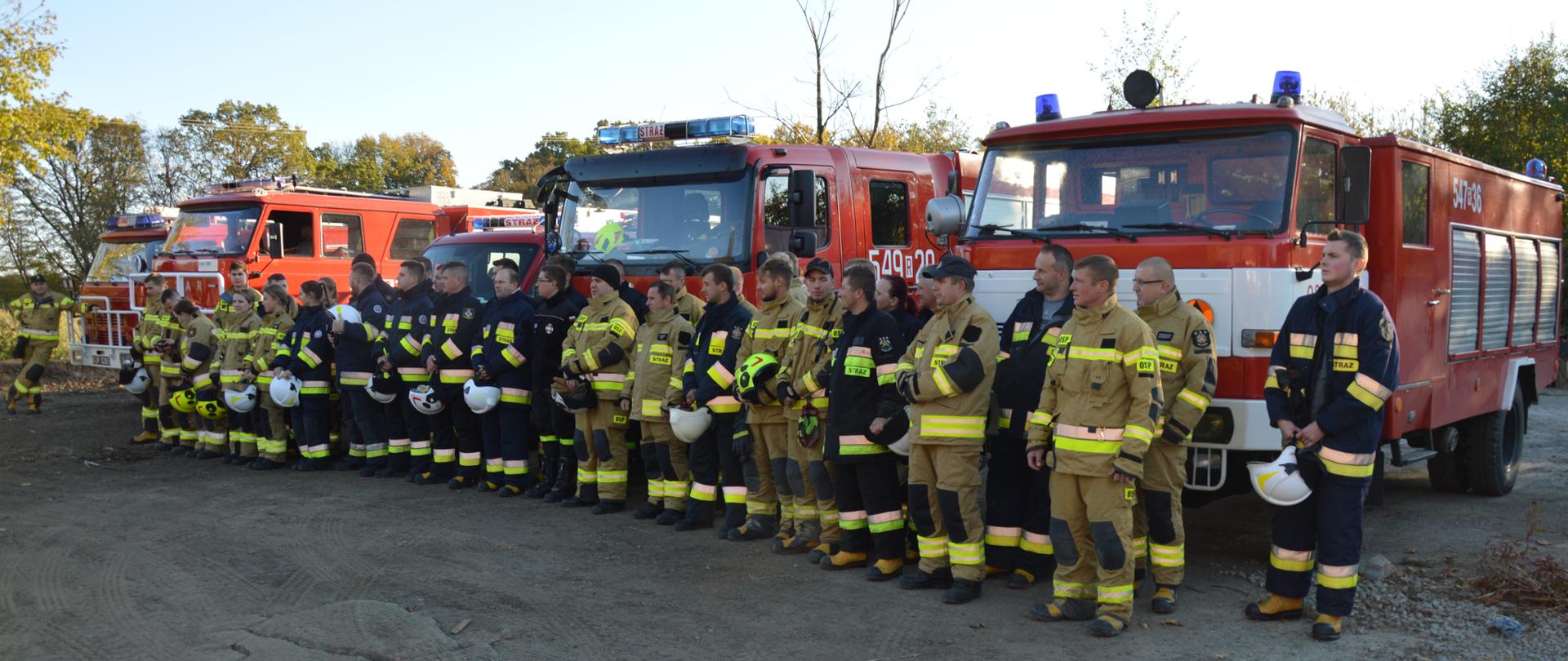 Zdjęcie przedstawia zbiórkę w dwuszeregu strażaków przed samochodami pożarniczymi. Strażacy ubrani w ubrania specjalne koloru piaskowego i granatowego. 