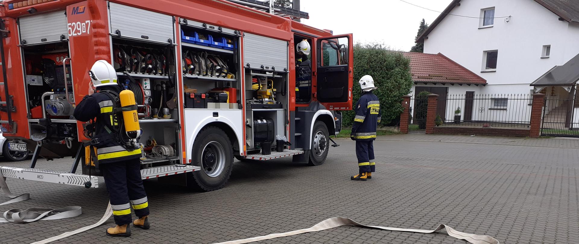 zdjęcie przedstawia strażaków ratowników Ochotniczej Straży Pożarnej podczas egzaminu praktycznego, na zdjęciu samochód ratowniczy z otwartymi skrytkami, rozwinięty wąż, dwóch strażaków podczas działań