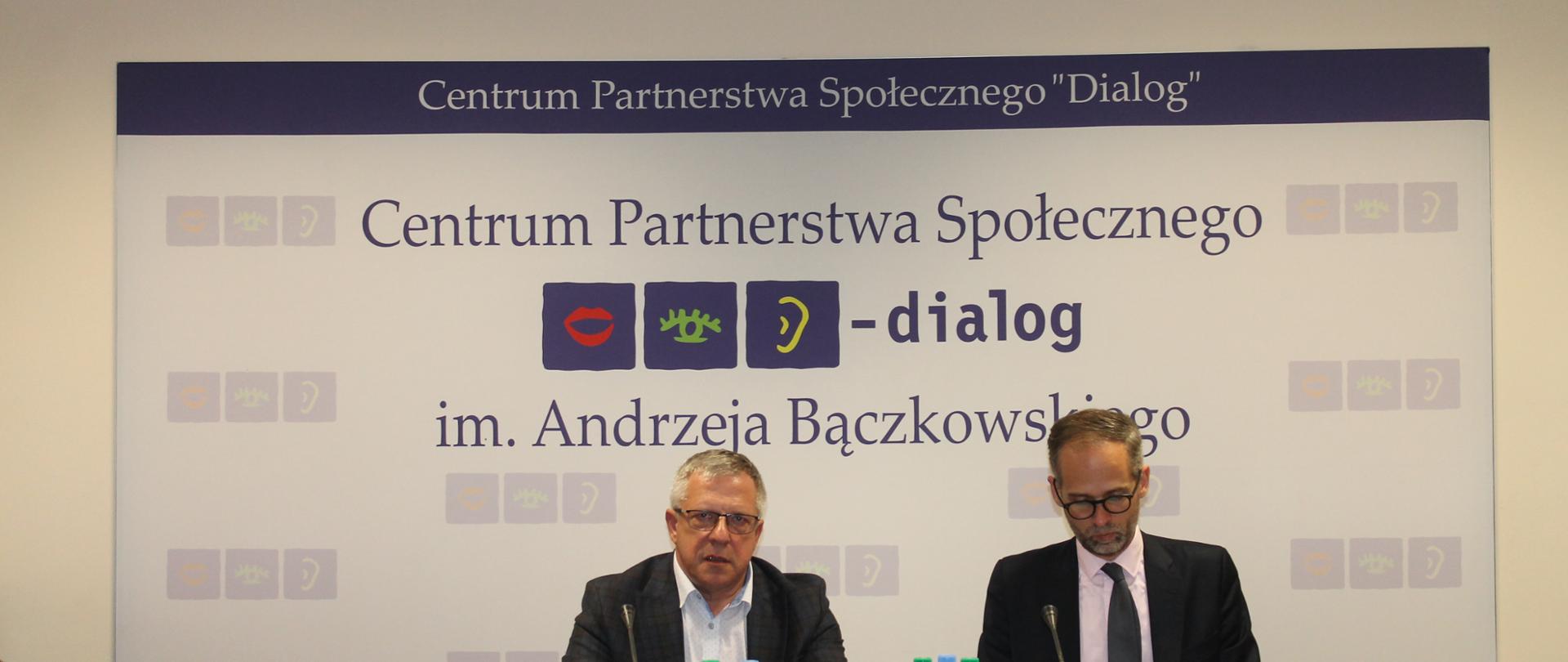 Zdjęcie przedstawia stół prezydialny za którym siedzą: Adam Guibourgé-Czetwertyński- Podsekretarz Stanu w Ministerstwie Klimatu i Środowiska, Leszek Miętek – Przewodniczący Zespołu. W tle siedzących znajduje się baner z napisem Centrum Partnerstwa Społecznego Dialog im. Andrzeja Bączkowskiego. Nad banerem widnieje Godło Polski.