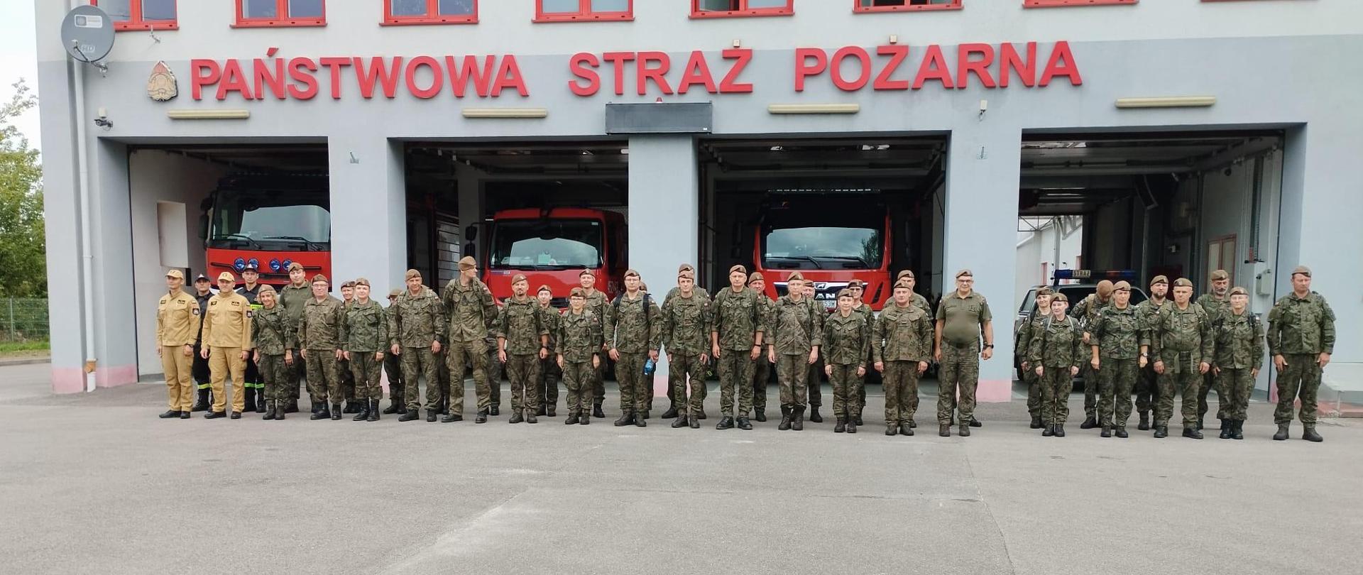 Na zdjęciu widoczni są funkcjonariusze Państwowej Straży Pożarnej oraz żołnierze Wojsk Obrony Terytorialnej, stojący w szyku na tle budynku Jednostki Ratowniczo-Gaśniczej nr 2 w Ostrowcu Świętokrzyskim. 