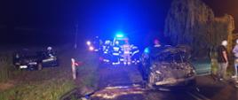Po lewej stronie w przydrożnym rowie stoi 1 z aut biorących udział w zdarzeniu. Po środku drogi stoi 2 samochód. Za nim stoją strażacy i karetka pogotowia ratunkowego