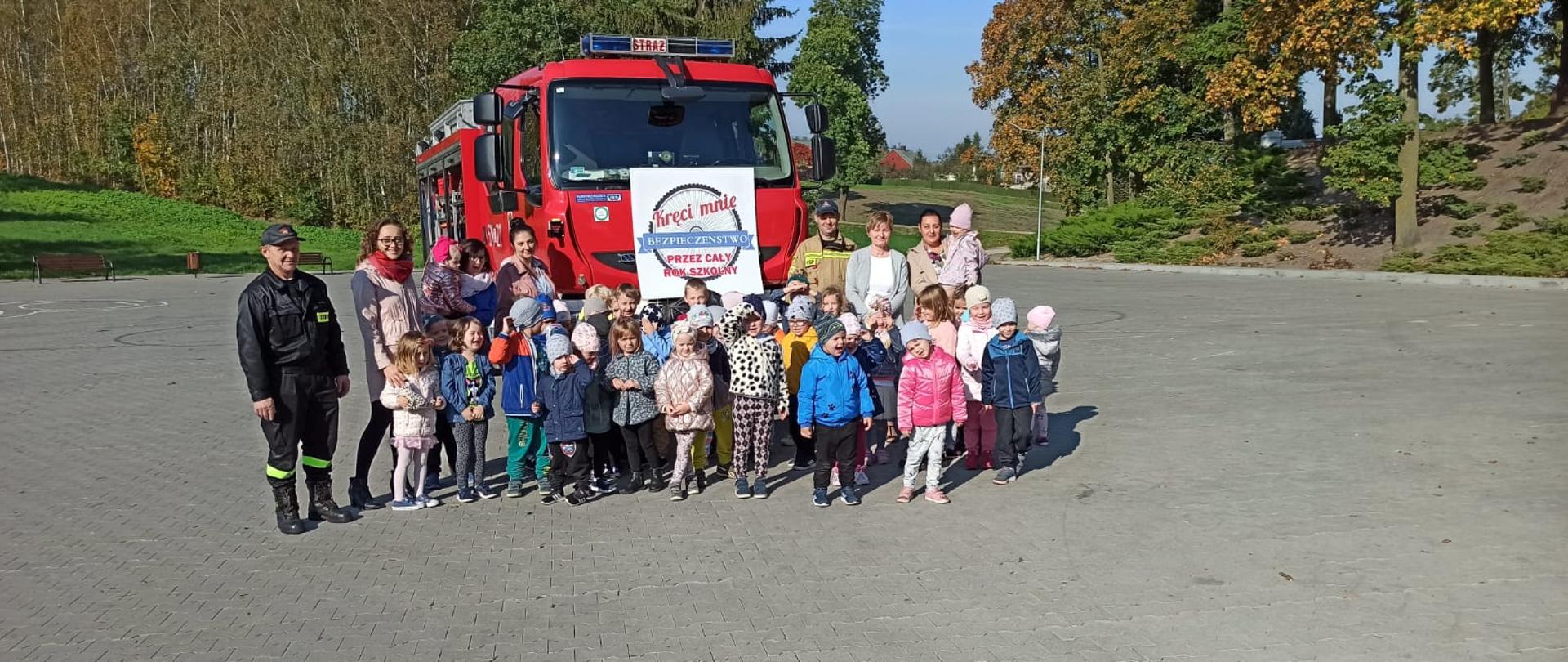 Zdjęcie przedstawia dzieci w wieku przedszkolnym, ich pięcioro opiekunów oraz dwóch strażaków. Wszyscy stoją w grupie na placu z kostki brukowej, za nimi szyld z hasłem kampanii prewencyjnej "Kręci mnie bezpieczeństwo przez cały rok szkolny", a w tle widać wóz strażacki oraz drzewa.