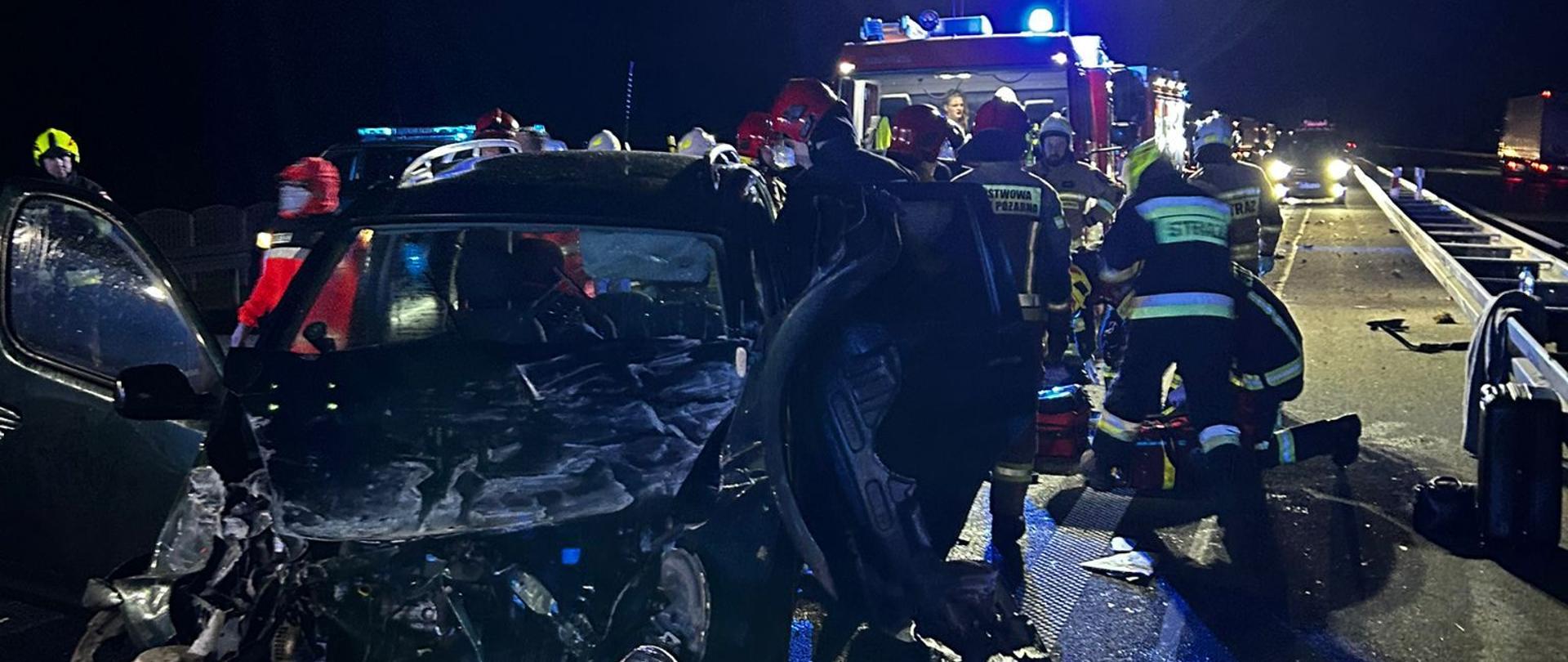Uszkodzony przód samochodu, w tle strażacy.