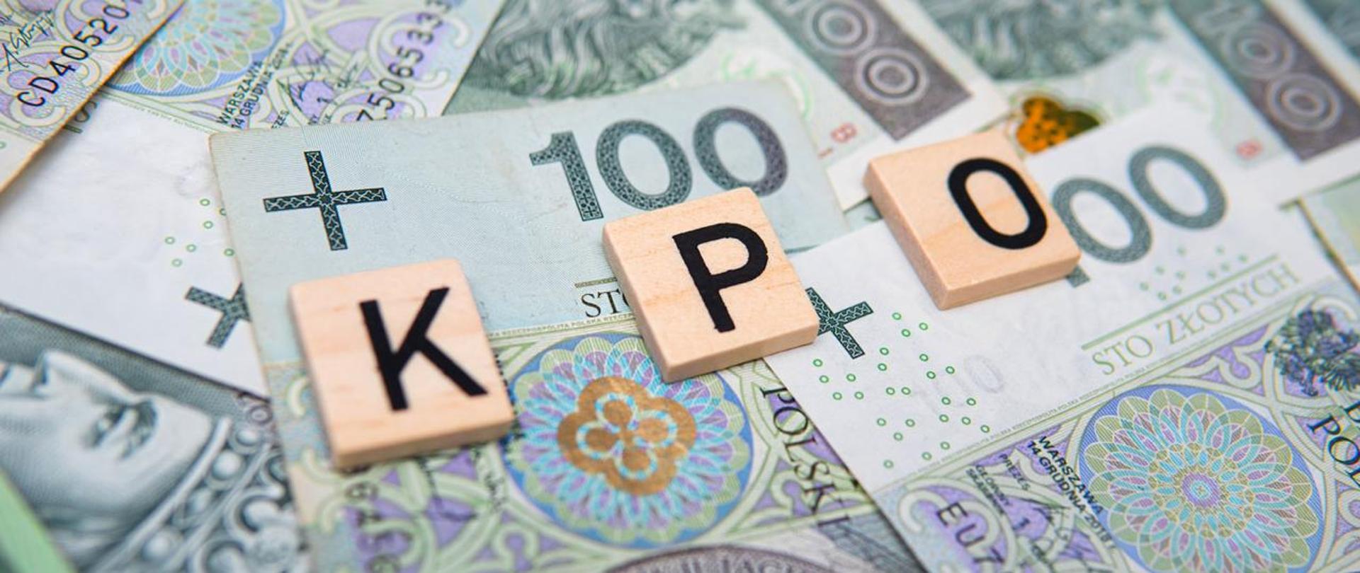 3 małe płytki drewniane z literami K, P, O na pliku banknotów stuzłotowych