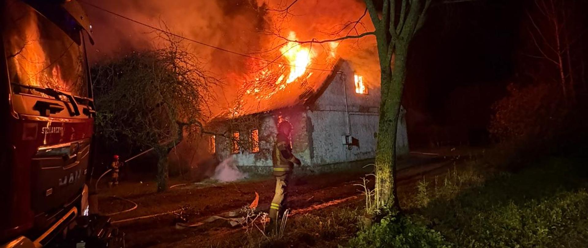 W pełni rozwinięty pożar budynku mieszkalnego. Ogień wydostaje się przez spaloną konstrukcję dachu. Strażak w trakcie rozwijania linii gaśniczej. Drugi strażak podaje prąd wody do wnętrza palącego się budynku. W lewej części zdjęcia widoczna jest również kabina samochodu pożarniczego.