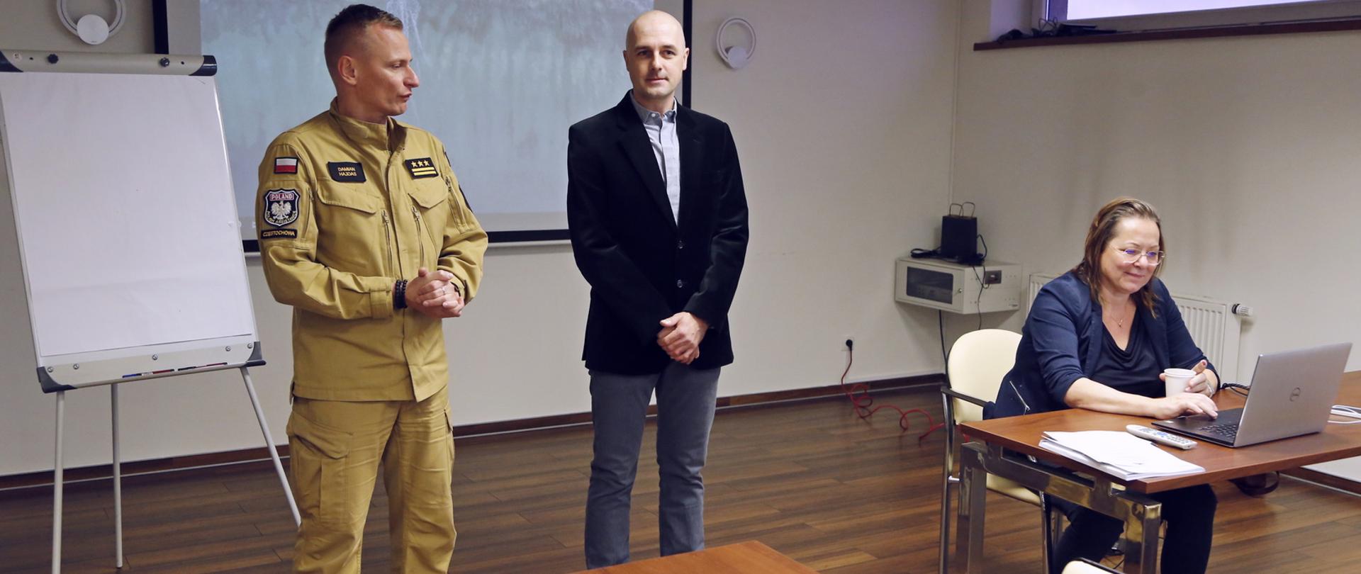 Od lewej: stojący Zastępca Komendanta Centralnej Szkoły PSP, mężczyzna w marynarce oraz siedząca przy stole kobieta obsługująca laptop