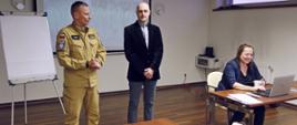 Od lewej: stojący Zastępca Komendanta Centralnej Szkoły PSP, mężczyzna w marynarce oraz siedząca przy stole kobieta obsługująca laptop
