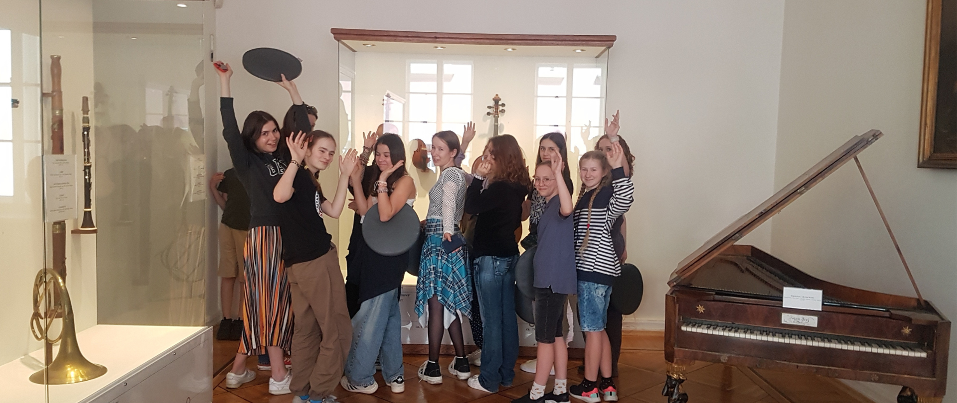 Grupa młodzieży stojąca w sali, w której z lewej strony stoi gablota z instrumentami, z prawej strony fortepian w kolorze brązowym.