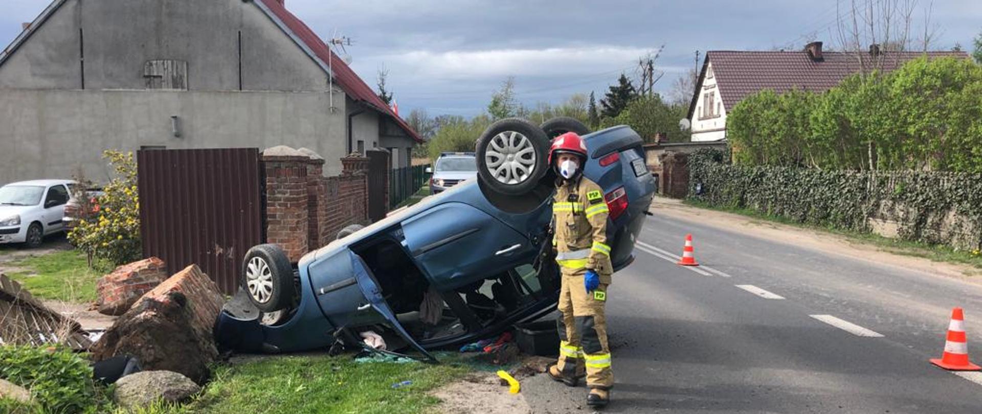 Na zdjęciu widać strażaka w bojowym mundurze, a w tle przewrócony na dach granatowy samochód osobowy.